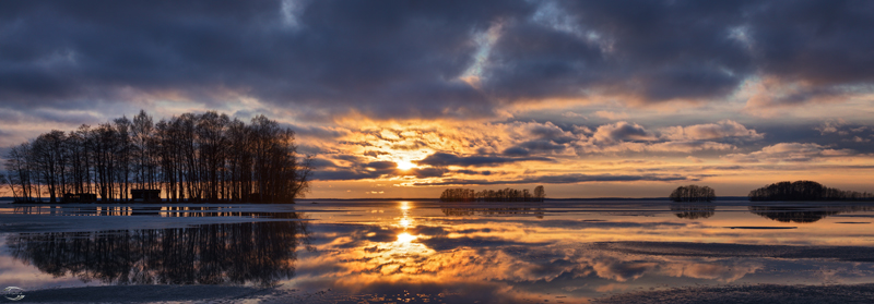 Panorama eines gefronenen Sees mit einem bewölkgem Himmel und der untegehenden Sonne am Horizont
