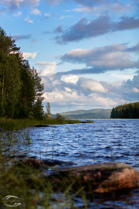 Blick über eine Wasserfläche entlang eines Waldes Richtung mehrerer Hügle