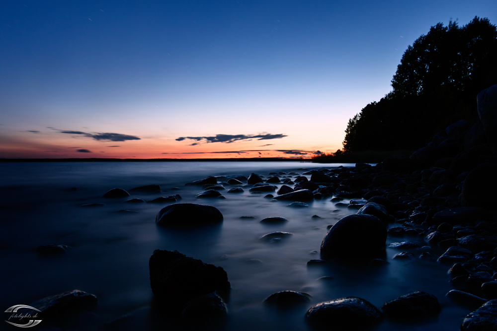 Blick über eine Wasserfläche zur Blauen Stunde mit Steinen im Vordergrund