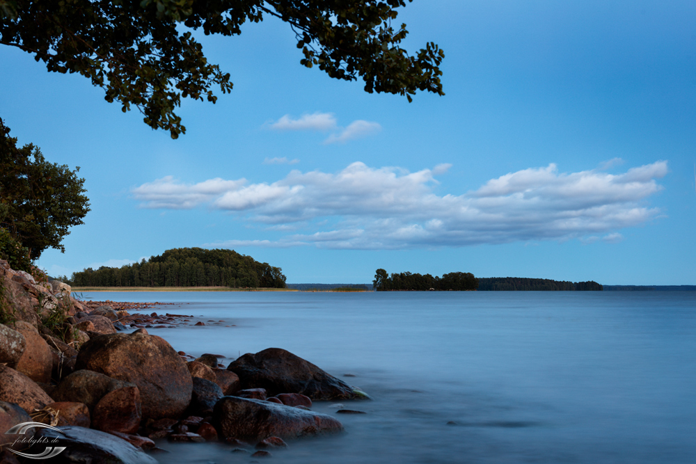 Blick über einen See Richtung zwei Inseln mit Steinen im Vordergrund links