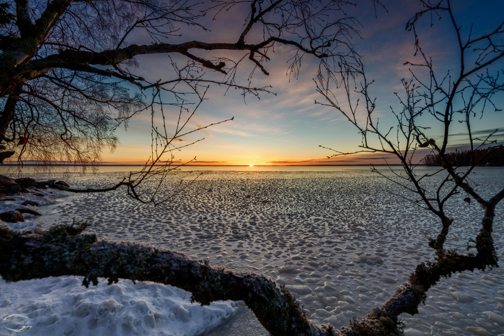 Blick über einen vereisten See vom Ufer aus mit der Sonne am Horizont