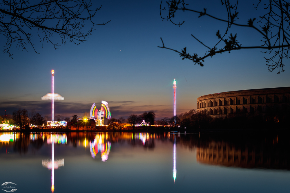 Blick über einen See auf Fahrgeeschäfte und das Nürnberger Kolloseum bei Nacht
